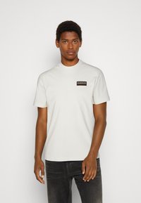 White cotton t-shirt with a crew neckline and short sleeves, featuring a small rectangular black and multicolored logo at the chest.