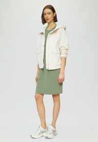 Leichte, kapuzenförmige cremefarbene Jacke mit elastischen Bündchen, kombiniert mit einem einfarbigen grünen Kleid. Das Model trägt graue und weiße Sneakers. Einfaches Design.