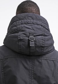 Cappotto imbottito nero con colletto alto e fibbia regolabile sul retro del cappuccio. Texture liscia con dettagli di cucitura visibili.