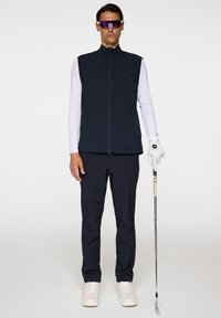 Marinblå golfväst över en vit långärmad skjorta, i kombination med marinblå byxor och vita sneakers. Accessoarer inkluderar lila solglasögon och en golfklubba.