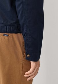 Giacca blu navy con polsini abbottonati, abbinata a pantaloni marrone chiaro. Il tessuto appare liscio con un design strutturato.