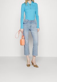 Polo de manga larga azul claro con cremallera, combinado con unos vaqueros recortados de lavado claro. Los accesorios incluyen un pequeño bolso naranja y zapatos marrones de tacón cuadrado.