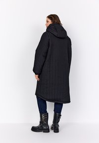 Nő, aki hosszú fekete steppelt kapucnis kabátot, sötétkék farmernadrágot és fekete csatos bőr csizmát visel, fehér háttér előtt áll.