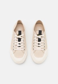 Beige canvas sneakers met witte rubberen zolen, voorzien van vlakke witte veters en een zwarte tab op de tong met een gele accent. Minimalistisch ontwerp.