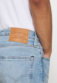 Acercamiento de una persona que lleva jeans Levi's de color azul claro con una etiqueta de cuero marrón de Levi Strauss & Co. y una mano en el bolsillo delantero.