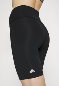 Fekete, elasztikus rövidnadrág sima anyagból. Magas derékkal rendelkezik, és a bal alsó lábszáron fehér Adidas logó található.