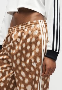 Άτομο που φοράει καφέ και λευκό παντελόνι Adidas με άσπρες ρίγες και κοντό λευκό μπουφάν με μαύρες ρίγες στο μανίκι.
