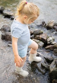 Enfant portant une chemise rayée bleu clair et un short en jean, marchant dans une eau peu profonde, avec des bottes en caoutchouc blanches, entouré de rochers.
