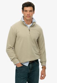 Beżowy sweter rozpinany z teksturowanej tkaniny, noszony na niebieskiej i białej koszuli w paski. Zestawiony z ciemnymi dżinsami.