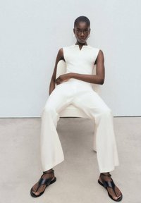 Jumpsuit bianco senza maniche con colletto, pantaloni a zampa, indossato con sandali neri. Modella seduta su una sedia di colore chiaro contro uno sfondo semplice.