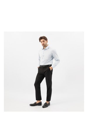 Mężczyzna stoi z rękami w kieszeniach, ubrany w jasnoniebieską koszulę, czarne spodnie, czarny pasek i czarne eleganckie buty na białym tle.