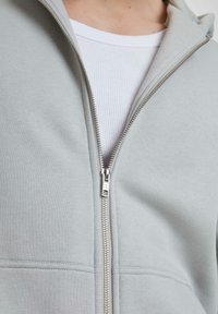 Hellgrauer Reißverschluss-Hoodie mit glatter Textur, ausgestattet mit einem silbernen Reißverschluss und zwei vorderen Taschen, über einem weißen gerippten Shirt getragen.