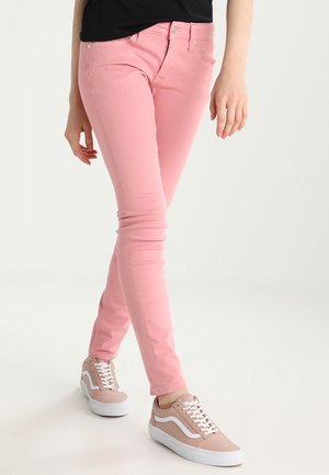 Vrouw draagt nauwsluitende roze spijkerbroek en lichtroze sneakers met witte veters, staand tegen een effen witte achtergrond.