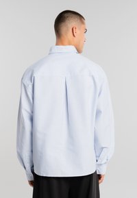 Hombre de espaldas vistiendo una camisa de botones a rayas en azul claro y blanco con mangas largas y pantalones negros, sobre un fondo liso.