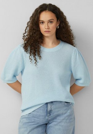 Jonge vrouw met lang krullend haar, gekleed in een lichtblauwe, gestructureerde trui met pofmouwen en een lichtblauwe spijkerbroek, staand tegen een effen achtergrond.