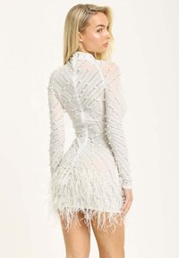 Short white dress with a fitted silhouette, made of sheer fabric adorned with pearls and sequins, featuring feather accents at the hem.