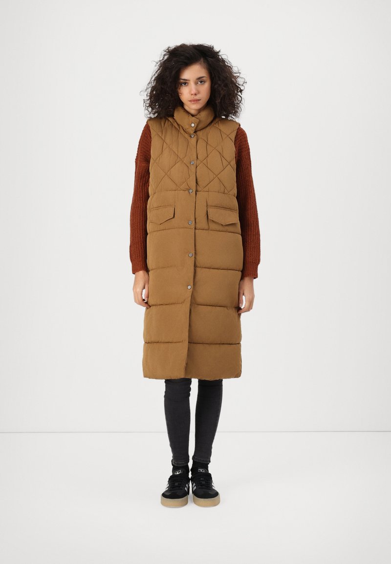 Gilet long matelassé marron avec un col montant, doté de deux poches avant, de boutons-pression, associé à un legging noir et des baskets.