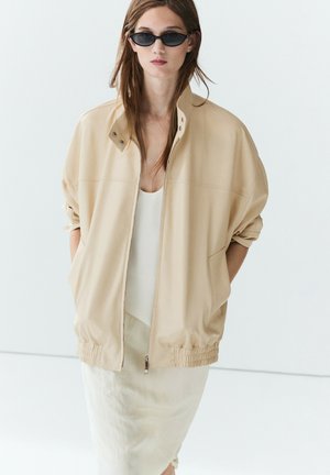 Frau mit schwarzen Cat-Eye-Sonnenbrillen, beiger Oversize-Zip-Up-Jacke und weißem Kleid, stehend vor einem schlichten weißen Hintergrund.