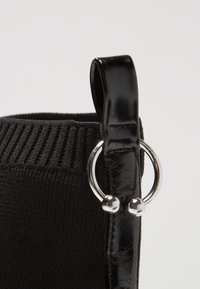 Bottine noire avec une tige en tissu texturé, des accents en cuir noir brillant et un détail en anneau argenté avec deux boules décoratives.