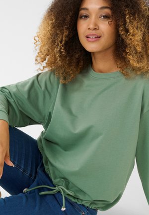 Sweat-shirt vert en matière douce avec un col rond, des manches longues et une taille ajustable par cordon. Associé à un jean bleu.