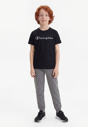 Barn med rødt hår iført en sort Champion t-shirt, grå joggingbukser og sorte sneakers, stående mod en hvid baggrund.