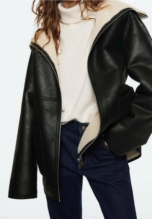Femme portant une veste en cuir noire avec doublure polaire crème, un pull à col roulé crème et un jean bleu foncé, main dans la poche.