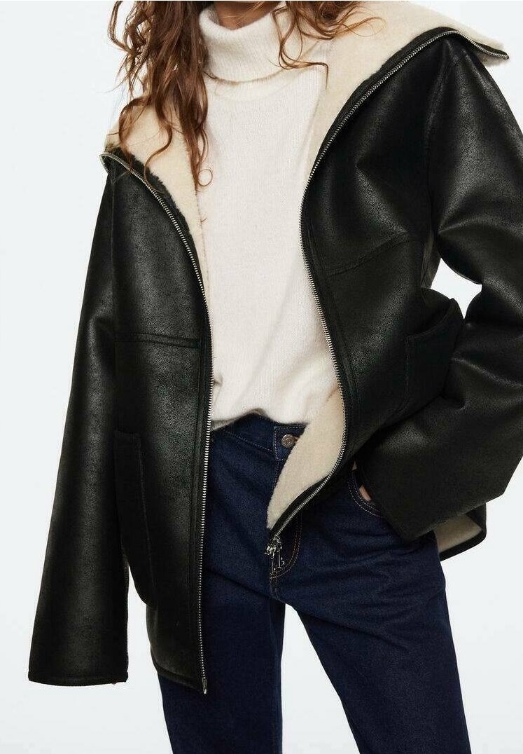 Femme portant une veste en cuir noire avec doublure polaire crème, un pull à col roulé crème et un jean bleu foncé, main dans la poche.
