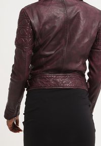 Giacca di pelle bordeaux con dettagli trapuntati sulle spalle, design aderente e superficie liscia, caratterizzata da una vita con cintura e hardware minimale.