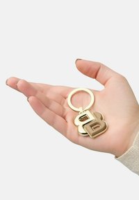 Gold keychain featuring a large stylized letter "B" with a brown accent, held in a hand against a plain background.