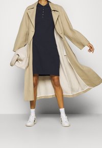 Beige trench coat with wide sleeves, worn over a dark navy polo dress. White sneakers with black accents. Textured knit accessory held under arm.