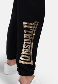 Black sweatpants with a gold textured logo printed vertically on the left leg. Features elastic cuffs at the ankles and a soft fabric texture.