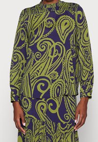 Nainen pukeutuneena yhteensopivaan pitkähihaiseen paitaan ja hameeseen, joissa on vihreitä paisley- ja pyörrekuvioita purppuranvärisellä taustalla.