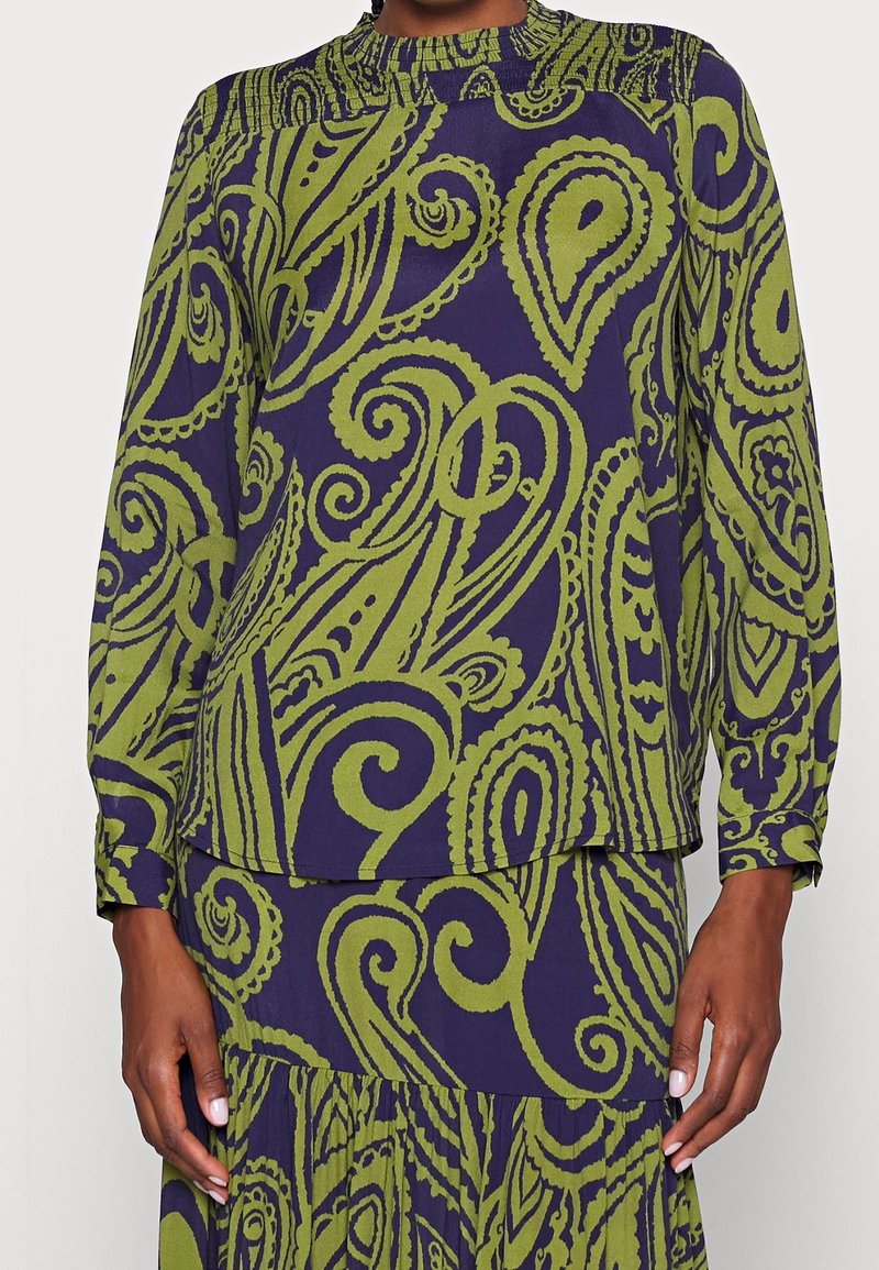 Nainen pukeutuneena yhteensopivaan pitkähihaiseen paitaan ja hameeseen, joissa on vihreitä paisley- ja pyörrekuvioita purppuranvärisellä taustalla.