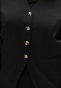 Fekete mellény testhezálló szabással és mély V-kivágással, öt díszes arany gombbal és két első zsebbel. Sima textúra.