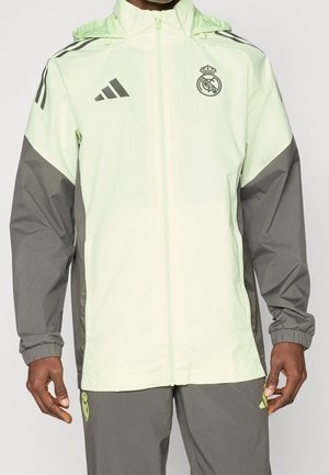 Mężczyzna ma na sobie jasnozieloną i szarą bluzę rozpinaną Real Madrid Adidas oraz pasujące szare spodnie z neonkowymi zielonymi detalami logo.