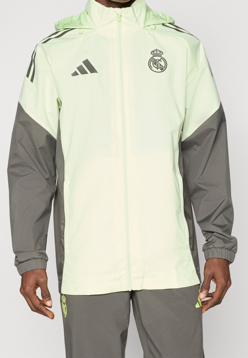 Un homme porte une veste zippée Real Madrid Adidas vert clair et gris ainsi qu'un pantalon gris assorti avec des détails de logo vert néon.