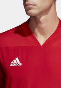 Maillot de sport rouge à col en V, en tissu lisse. Présente un logo Adidas blanc sur le côté gauche de la poitrine et des accents texturés aux épaules.