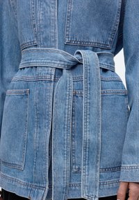 Hellblaue Jeansjacke mit großen Fronttaschen und einem gebundenen Taillengürtel, getragen von einer Person, die von der Mitte des Torsos bis zur Hüfte zu sehen ist.