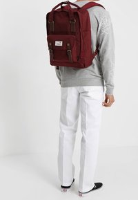 Bordowy plecak z materiału z górnym uchwytem, regulowanymi szelkami, przednią kieszenią z klapką i metalowymi elementami. Noszony przez osobę w szaro-białym stroju.