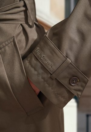 Khakifarbener Trenchcoat-Mantel mit den Stickereien "French Connection" an den Ärmeln, ausgestattet mit einem Knopfverschluss und einer glatten Textur.