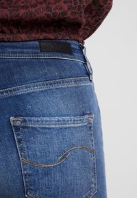 Jeans de mezclilla azul oscuro con un diseño de bolsillo trasero bordado, que presenta una etiqueta negra en la cinturilla y una textura suave.