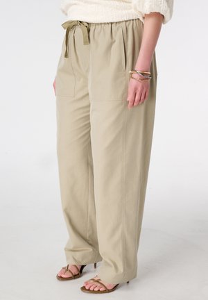 Pantalon beige à jambes larges avec poches avant et taille à cordon, porté avec des sandales à talons marron à lanières et un pull crème.