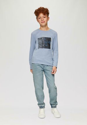 Chemise à manches longues bleu clair avec un imprimé graphique et du texte, associée à un jean en denim clair et des baskets blanches à semelle texturée.