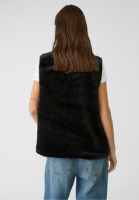 Zwart nepbontvest met een zachte textuur, voorzien van een hoge hals en een open voorkant. Draagt over een wit T-shirt en gecombineerd met lichtblauwe jeans.