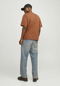 Brun kortærmet T-shirt parret med lyseblå løstsiddende jeans. Sort kasket båret baglæns; detaljer inkluderer subtil syning og mørke sko.