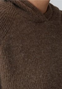 Zbliżenie na osobę w grubym, brązowym, puszystym swetrze z wysokim, zaokrąglonym dekoltem.