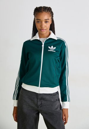 Mørkegrøn zip-up jakke med hvid krave og manchetter. Har hvid Adidas-logo og tre striber ned ad ærmerne. Blødt stoftekstur.