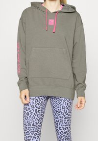 Sweat à capuche vert olive avec cordons de couleur rose, poche avant et logo Nike rose sur la manche. Porté avec des leggings imprimé léopard violet.