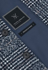 Wolblendstof met een blauw-wit patroon, voorzien van een zwart label en knoop. Op het label staat: "PAKO LORENTE" en "PREMIUM WOOL."