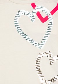 Maglietta beige con due cuori scintillanti in bianco e argento con motivi alternati, accentuata da una striscia diagonale rosa.